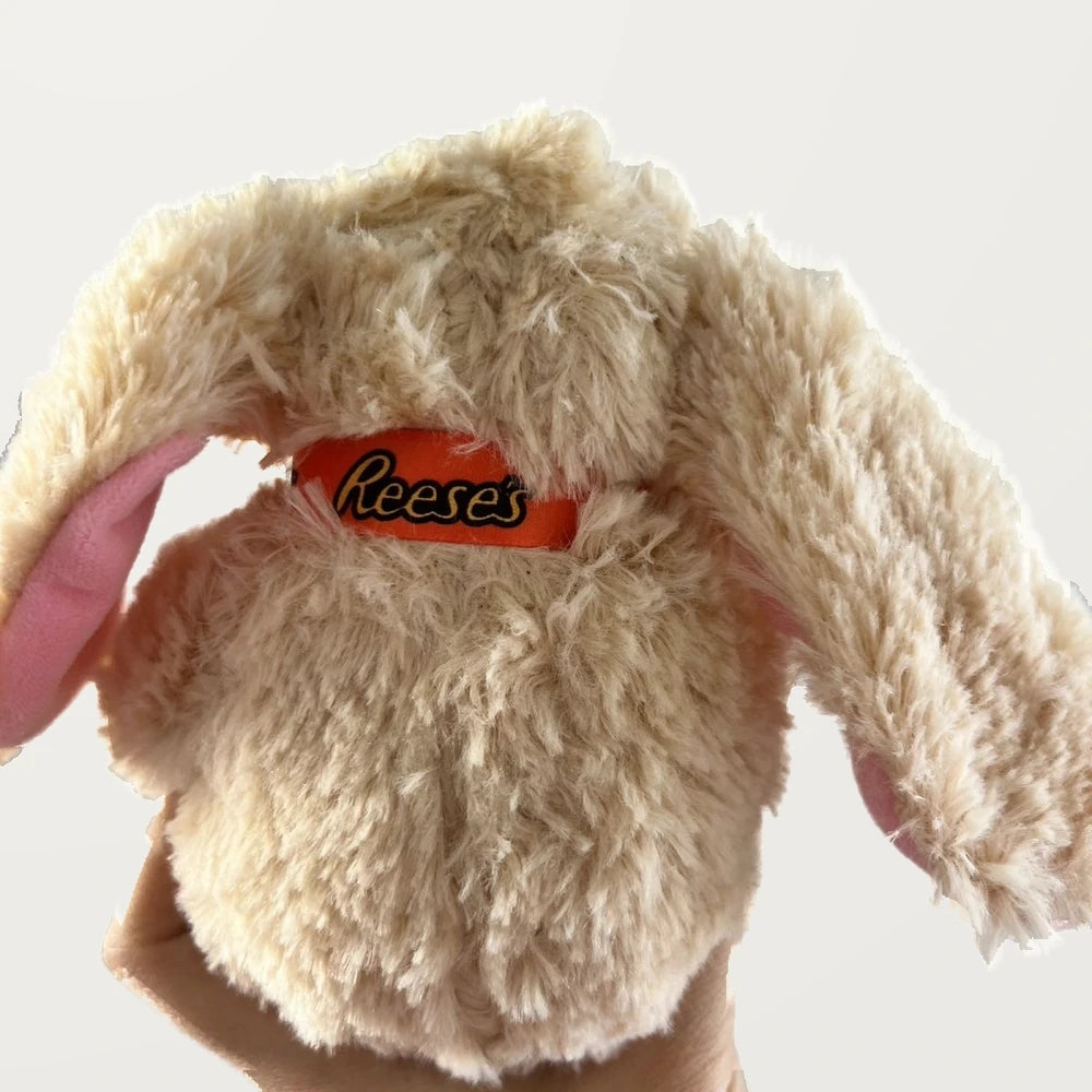 Back view of a beige floppy-ear bunny plush showing its textured fur, pink ear lining, and orange neck ribbon.