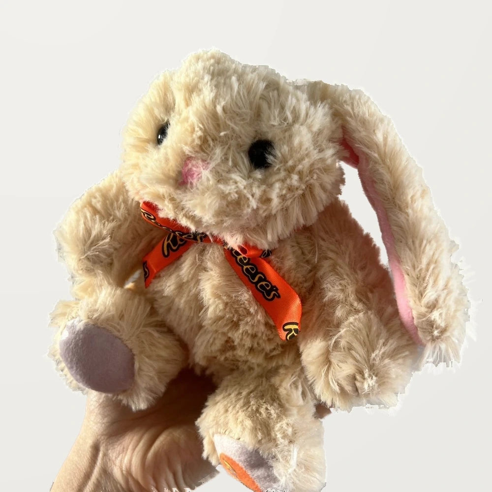 Three-quarter view of a beige bunny plush showing pink-lined ears, an orange bow, and lavender-gray foot pads.