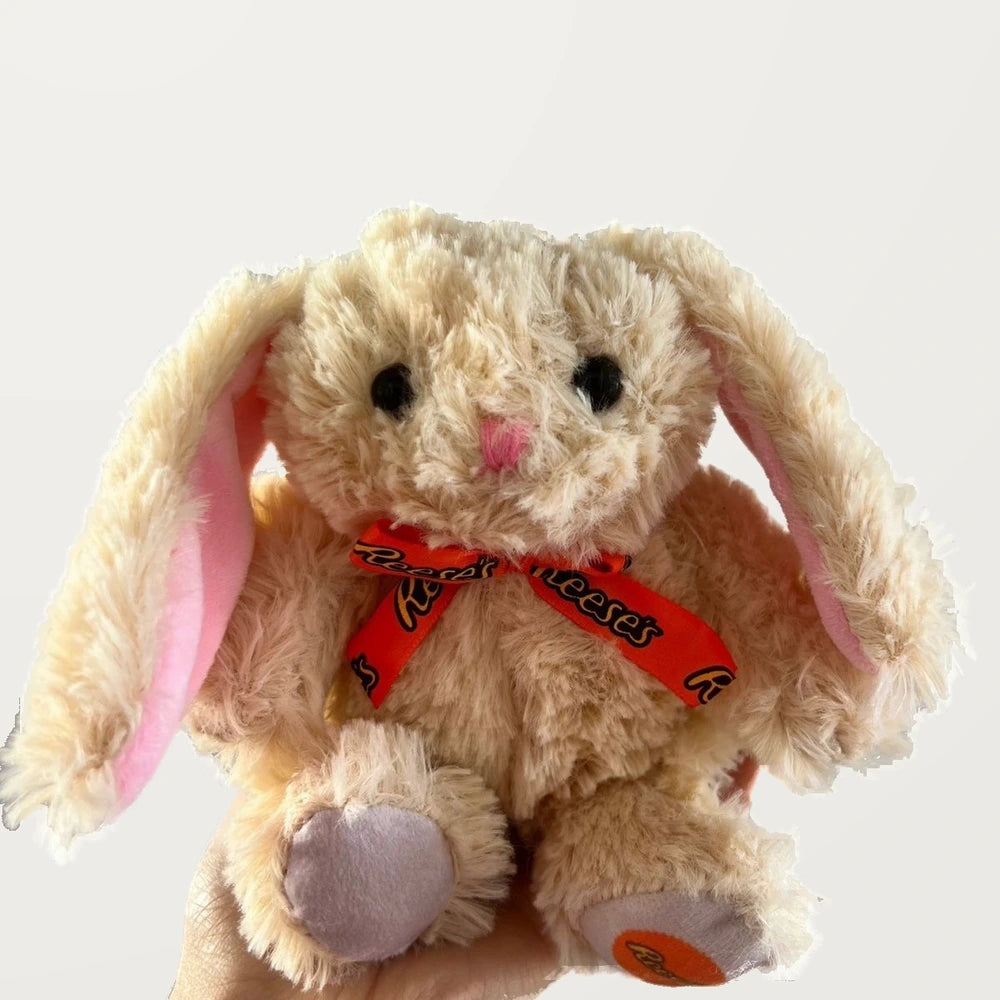 Close-up front view of a beige bunny plush showing its black eyes, pink embroidered nose, and orange printed bow.