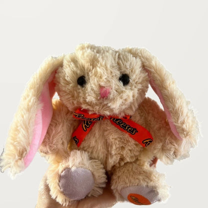 Close-up front view of a beige bunny plush showing its black eyes, pink embroidered nose, and orange printed bow.