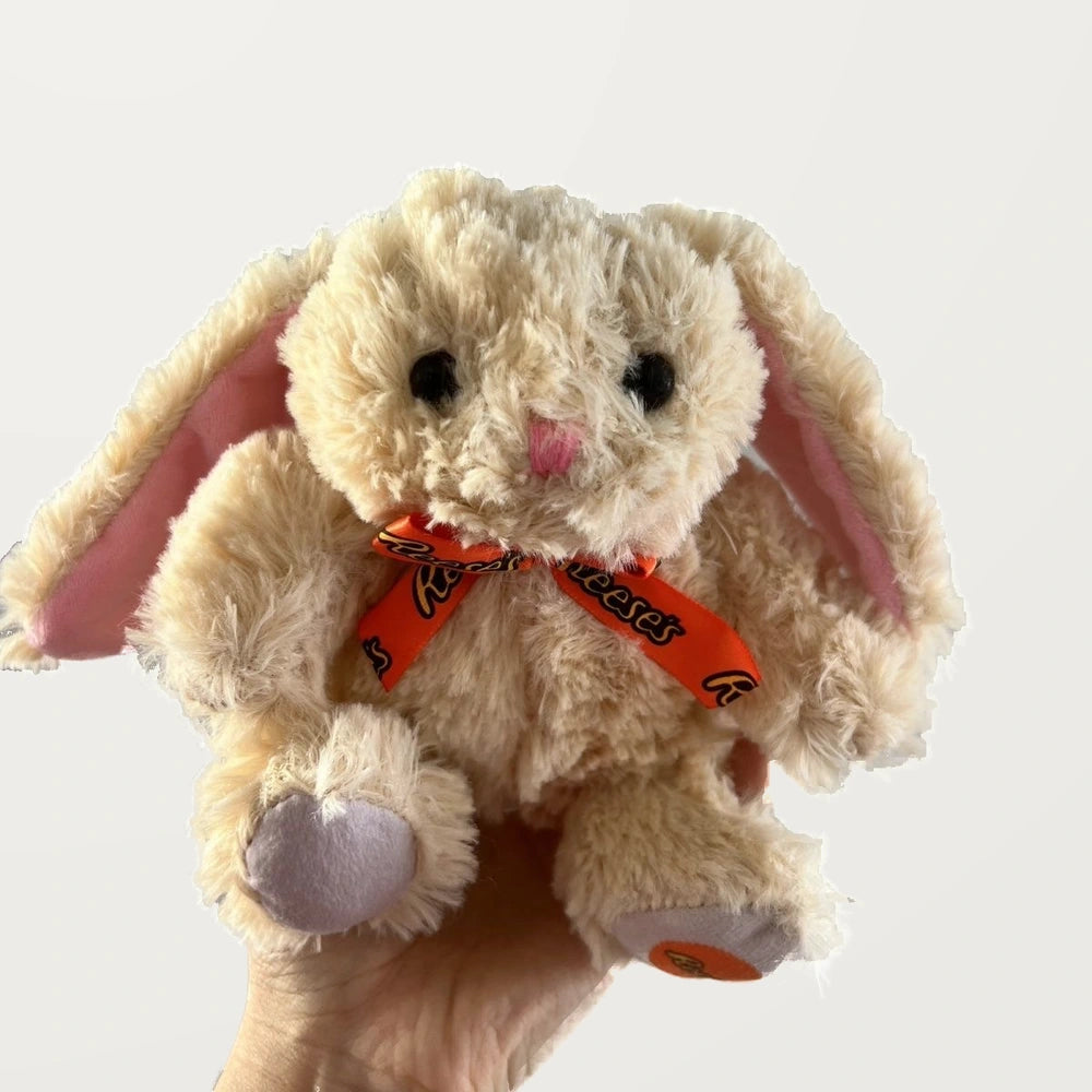 Front view of a beige floppy-ear bunny plush with a pink nose, orange bow, and lavender-gray foot pads.