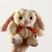 Front view of a beige floppy-ear bunny plush with a pink nose, orange bow, and lavender-gray foot pads.