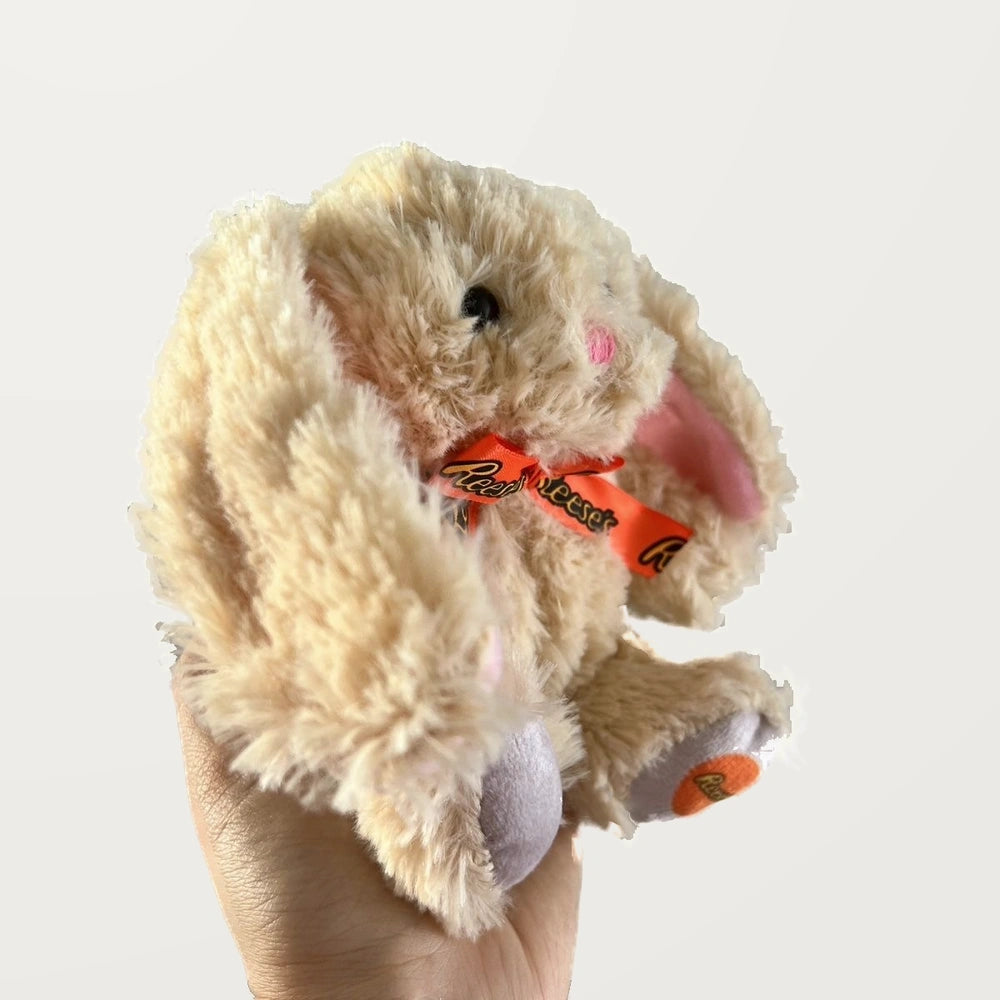 Side view of a beige floppy-ear bunny plush with an orange bow, held in one hand.