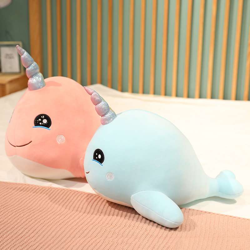 Two narwhal plushies, one pink and one blue, sitting on a bed. 