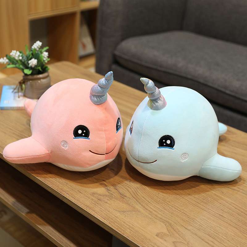 Two narwhal plushies, one pink and one blue, sitting on a bed. 