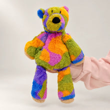 Rainbow bear plush with a black nose and long floppy legs, supported by an adult hand.
